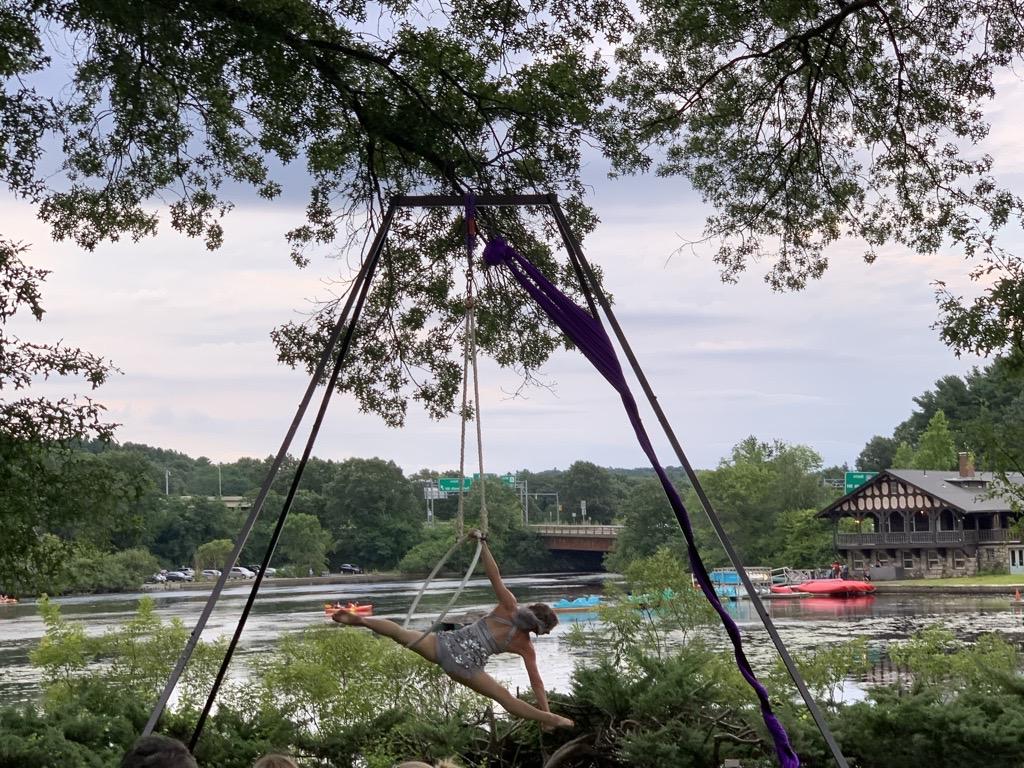 Performer doing a split in the air, suspended by a large ring tied to a support. In the background is a river view with bright colored boats and, in the distance, an old-fashioned single-story wooden structure.